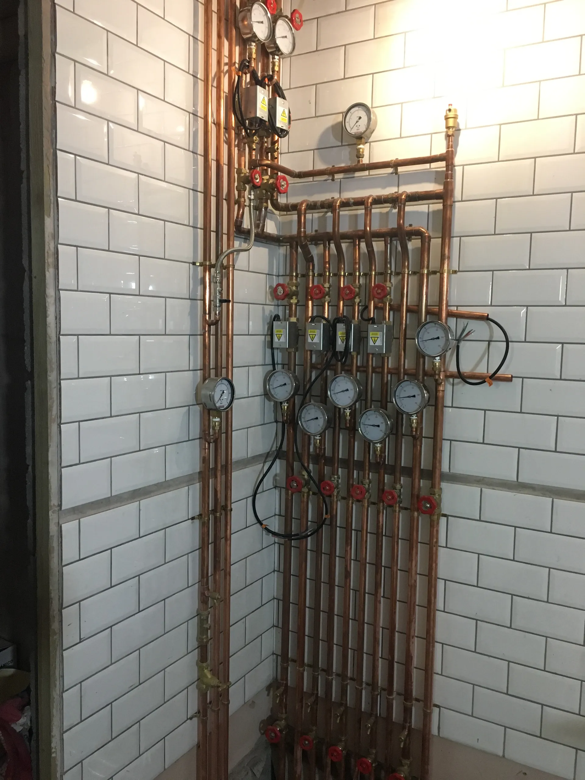 Elaborate copper pipework manifold with numerous pressure and temperature dial gauges and red gate valves on a white metro-tiled wall.