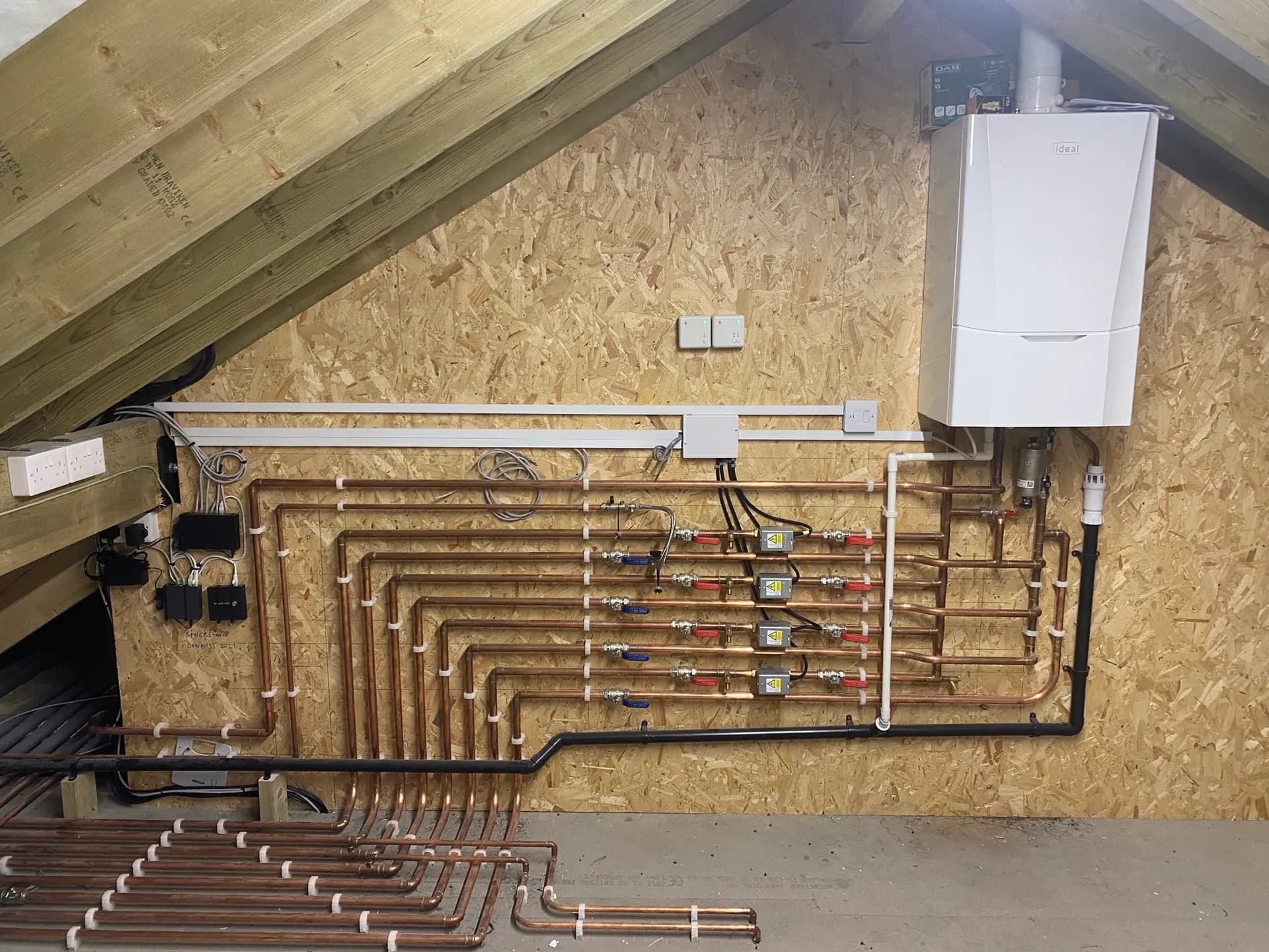 Loft mechanical install with a wall-hung combi boiler, soldered copper pipework, zone valves and copper underfloor-heating loops across the joists.