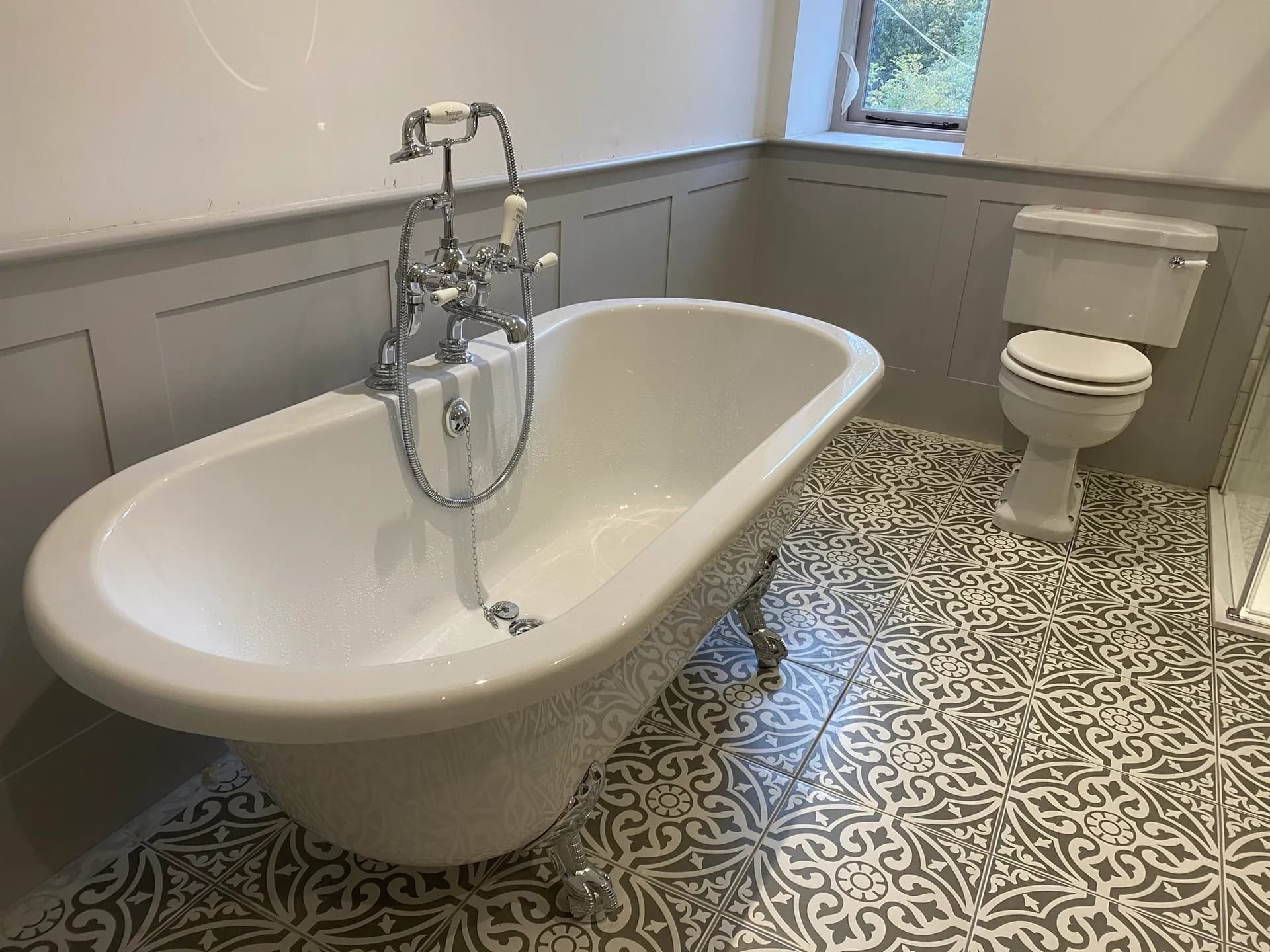 White roll-top clawfoot bath with a chrome telephone-style bath and shower mixer on grey-and-white patterned floor tiles.